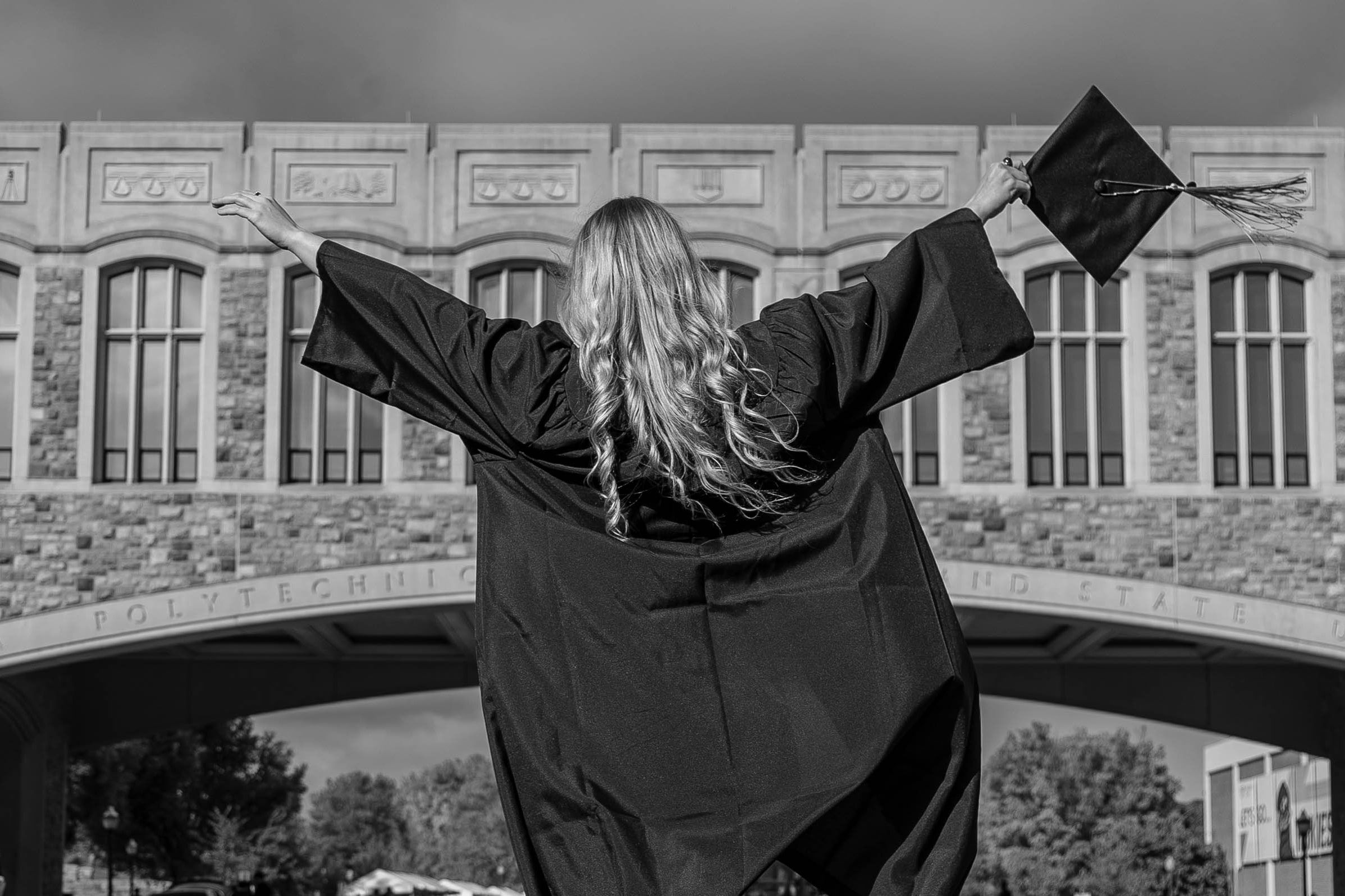 Graduate in cap and gown with arms open in front of a campus arch, in black and white